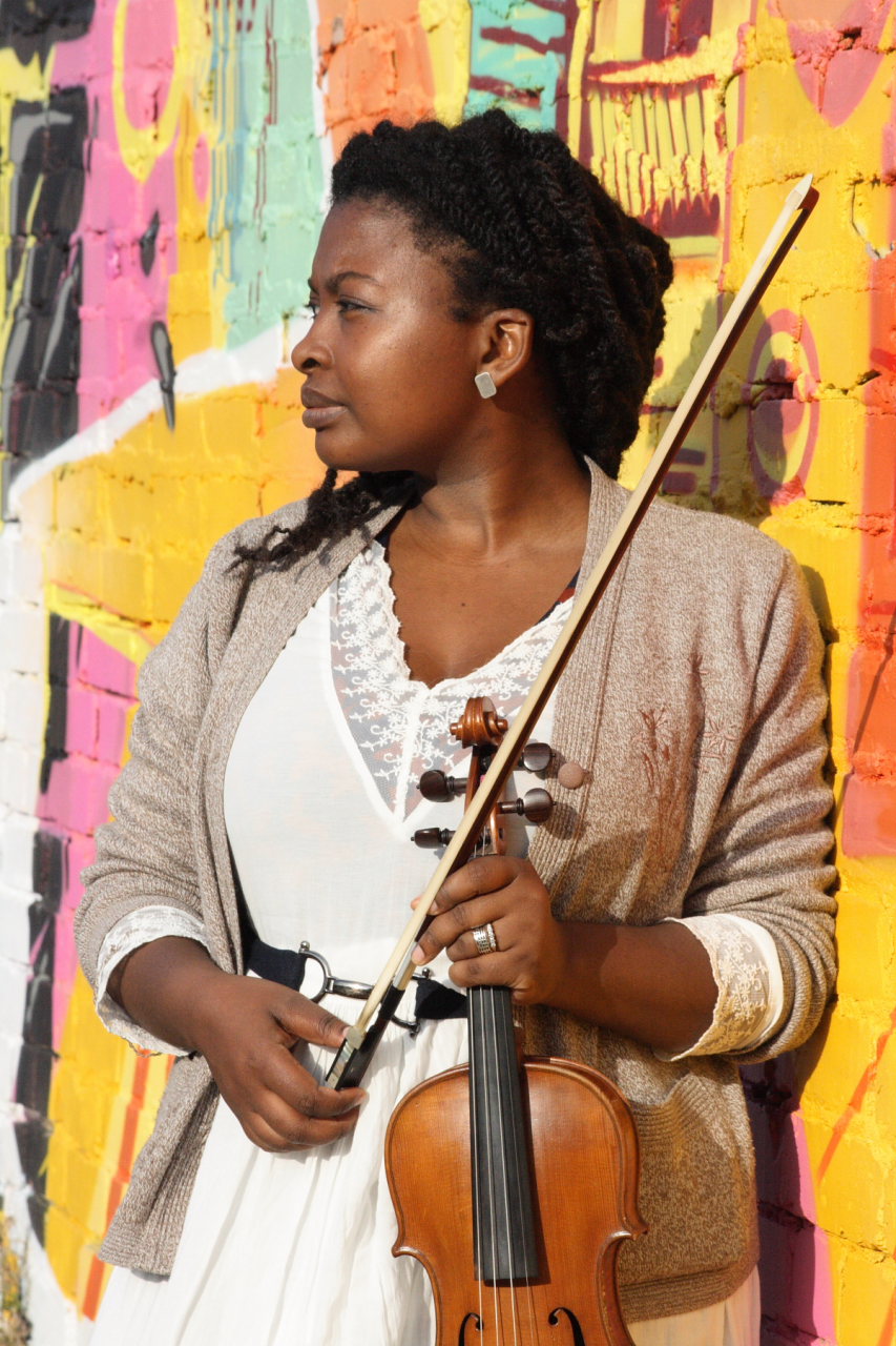 Portrait of Germa Adan standing against a colourful brick wall holding her violin